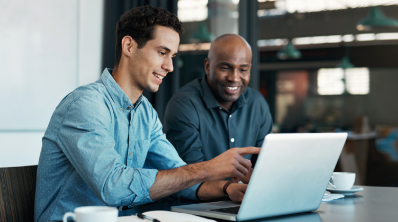 Two men are sitting in from of a laptop, both with a smile on their face. The man on the left is pointing at the screen.